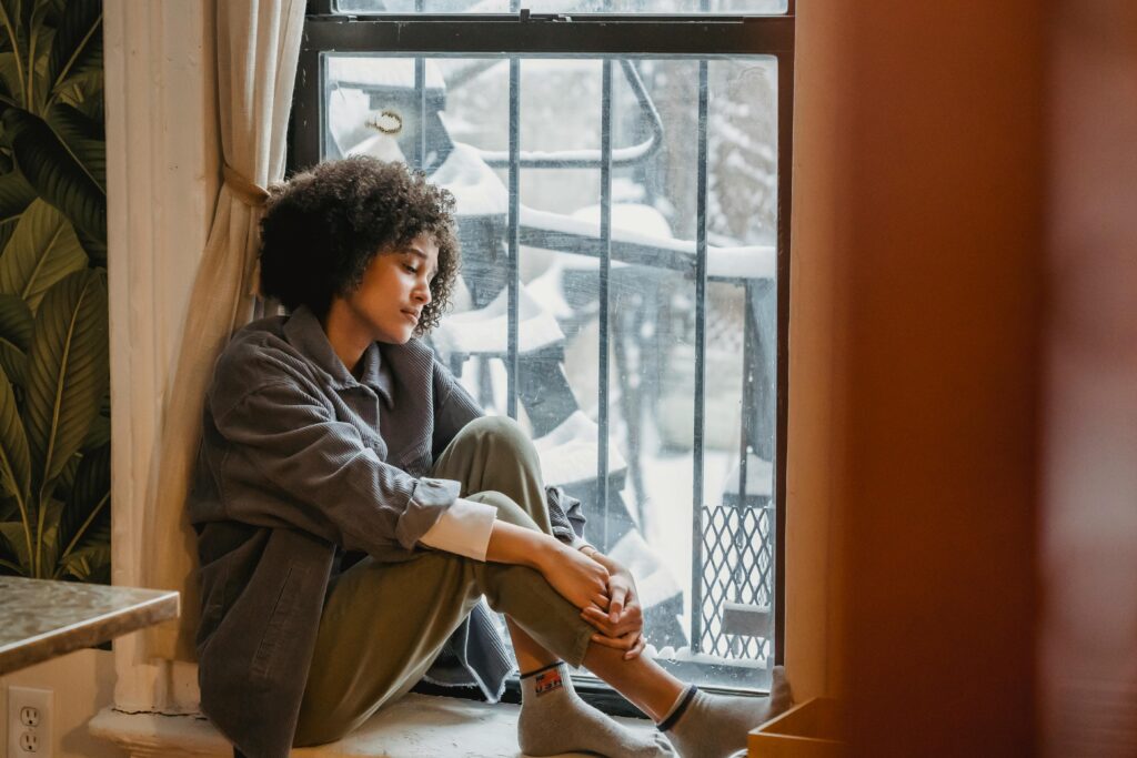 Side view full body of upset African American woman embracing knees while sitting in solitude near window and looking down