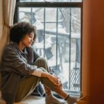 Side view full body of upset African American woman embracing knees while sitting in solitude near window and looking down