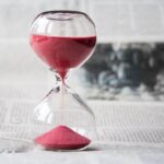 Close-up of a transparent hourglass with pink sand flowing, placed on a newspaper background.