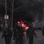 Riot police in gear confronting a street fire during an urban protest at night.