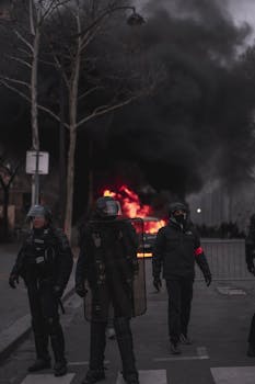 Riot police in gear confronting a street fire during an urban protest at night.