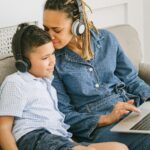 A mother and son sitting on a sofa, both with headphones, sharing a moment while using a laptop.