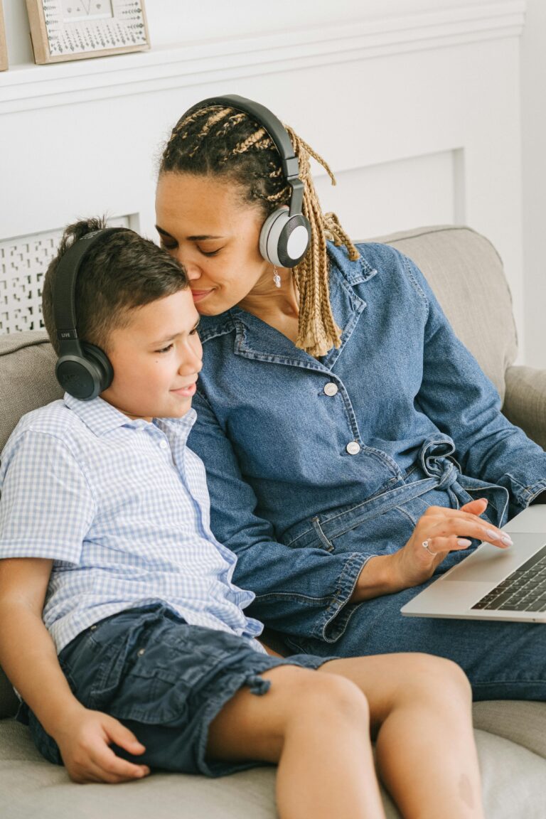 A mother and son sitting on a sofa, both with headphones, sharing a moment while using a laptop.