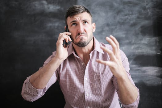 Adult man in pink dress shirt looking stressed while talking on a smartphone indoors.