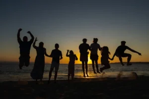 A group of friends or family on the beach during sunset, jumping up in unison, some holding hands.