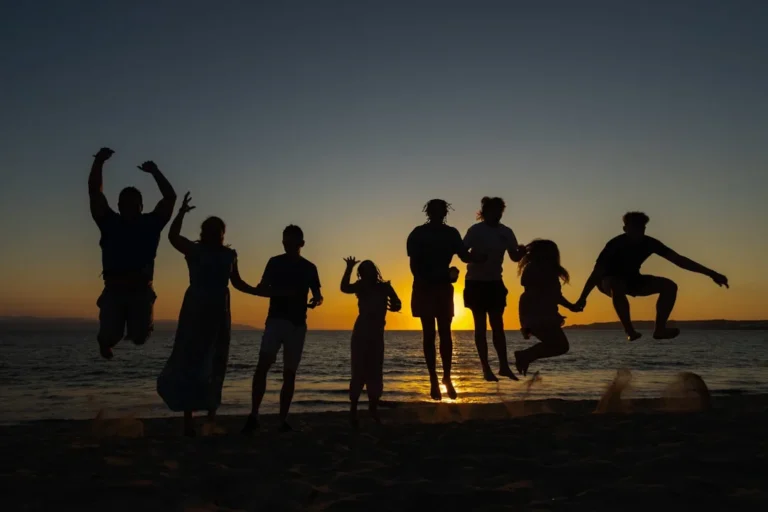 A group of friends or family on the beach during sunset, jumping up in unison, some holding hands.