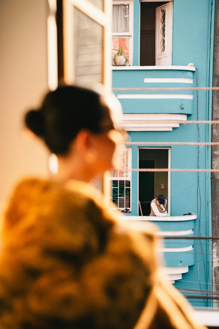 A woman gazes from a window, observing a person in a neighboring balcony, bathed in warm light.