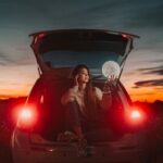 A woman seated in a car trunk holding a glowing moon against a vibrant sunset backdrop.