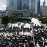 A large group of people with umbrellas gather on a rainy city street, surrounded by skyscrapers.