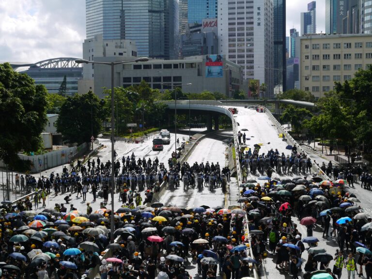A large group of people with umbrellas gather on a rainy city street, surrounded by skyscrapers.