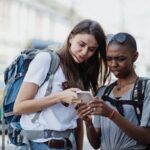 Two young women with backpacks consult a smartphone while exploring an urban area on a sunny day.