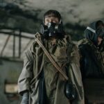 Two people in gas masks explore an abandoned, dystopian environment in survival gear.