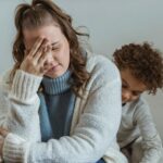 Frustrated mother with hand on forehead and closed eyes sitting near African American son near wall in room at home