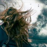 A woman with disheveled hair caught in strong wind against a stormy backdrop, capturing motion and chaos.