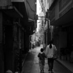 A mother and child walking hand in hand down a narrow city alleyway in black and white.