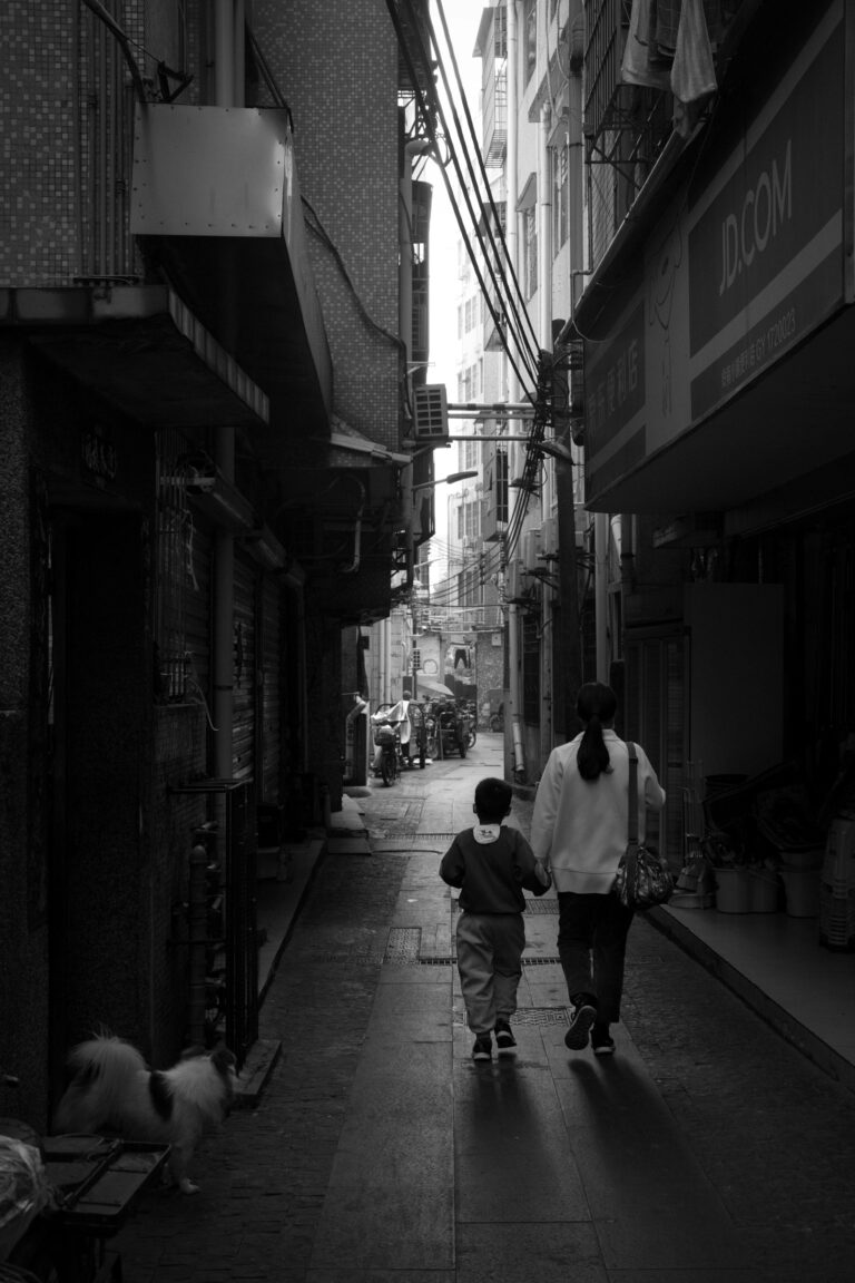 A mother and child walking hand in hand down a narrow city alleyway in black and white.