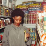 A smiling woman examines products in a colorful grocery store aisle.