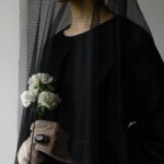 A woman wearing a black veil holds white flowers in a solemn, indoor setting.