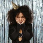 African American woman looking sad at a party, wearing a black outfit and party hat.