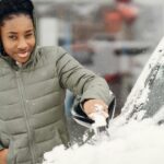 A young woman wearing a jacket clears snow from her car during a winter day.