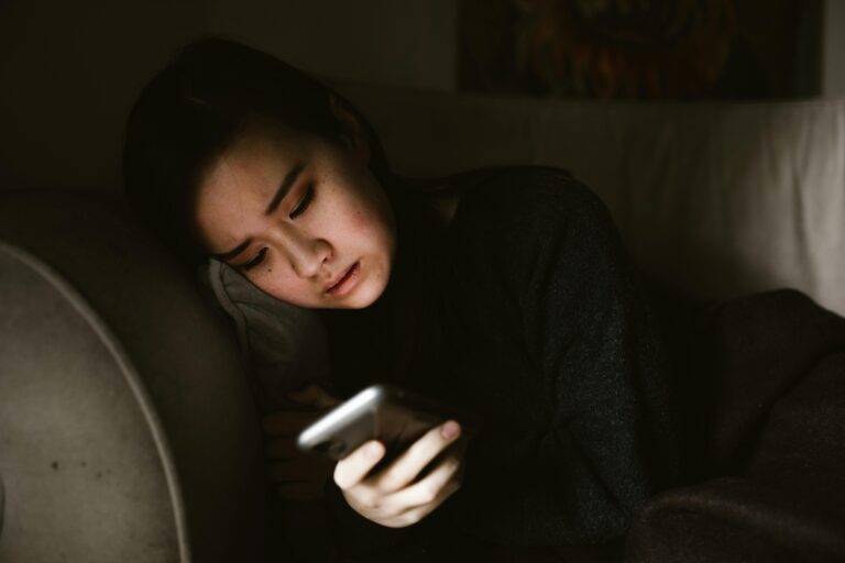 A young woman lying on a couch looking at her smartphone in a dimly lit room.